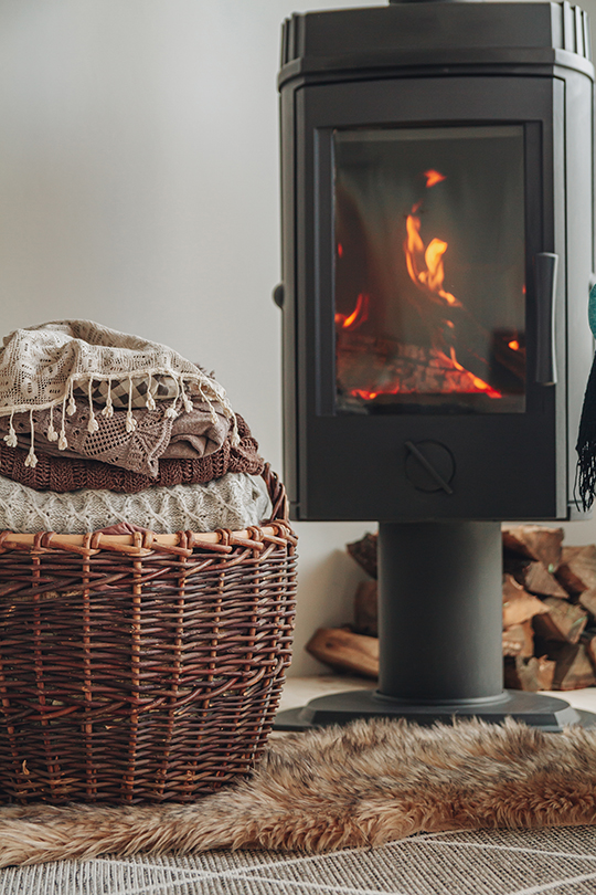 A stack of warm clothes in a wicker basket near an iron fireplace with a glass door