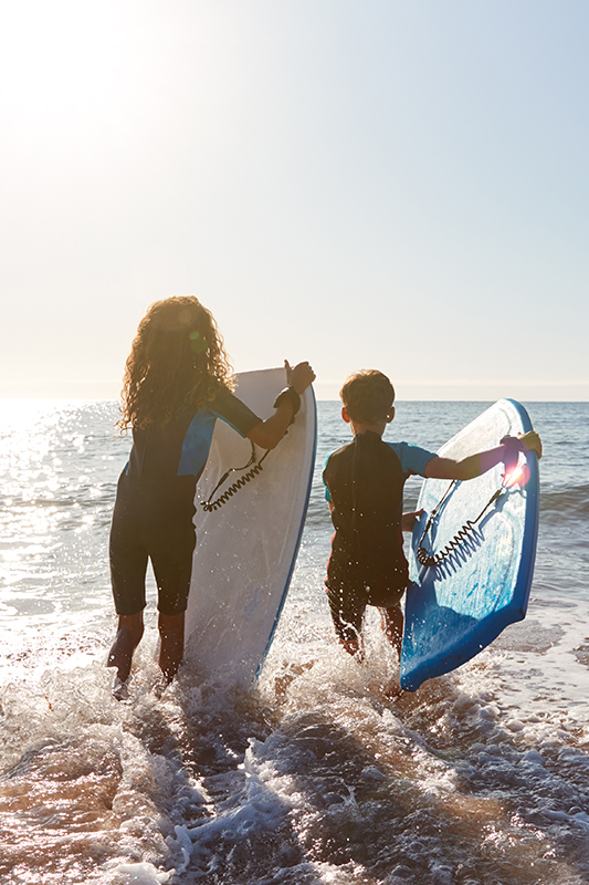 Two children wearing wetsuits carrying bodyboards into the sea at the beach